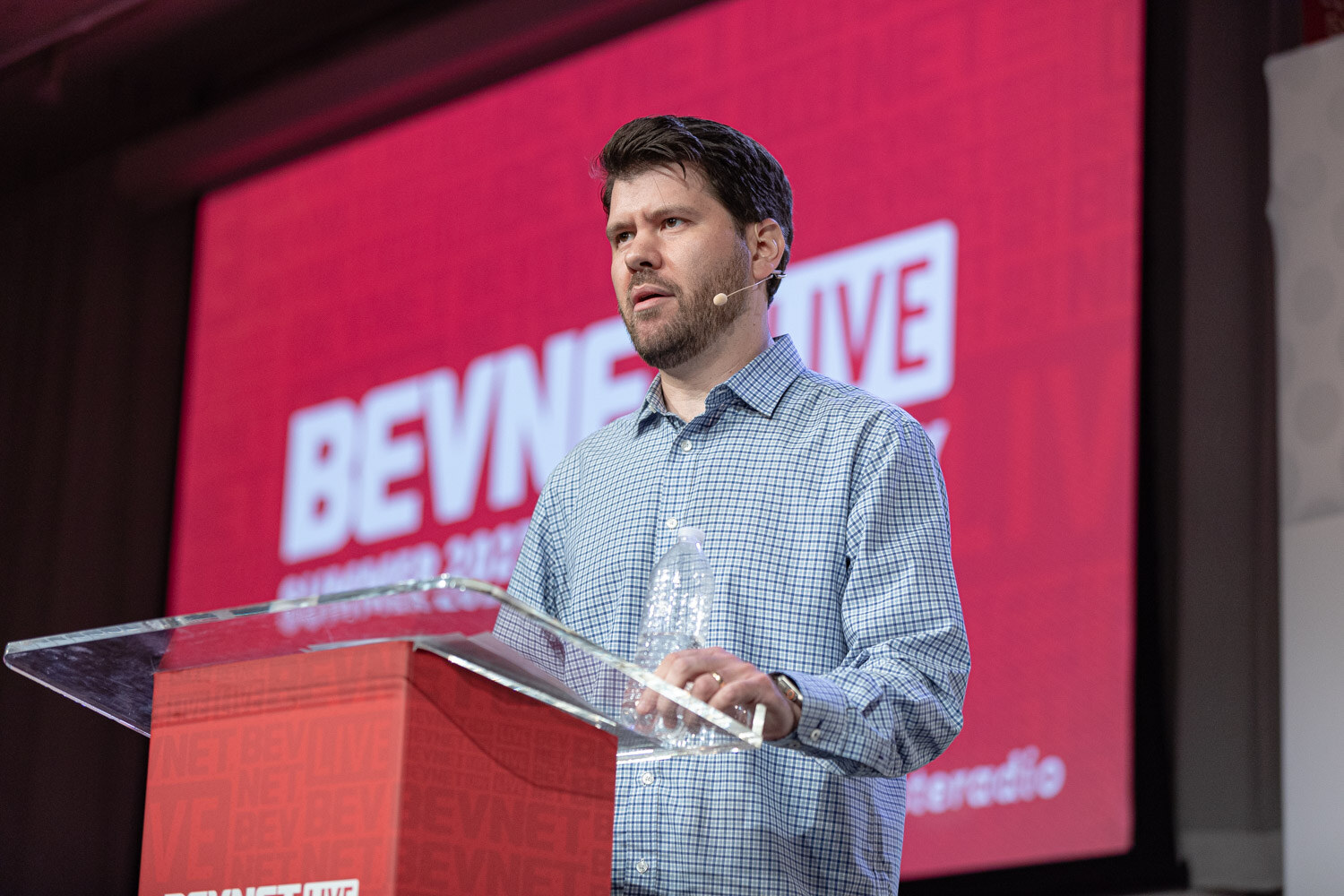 A man in a blue checked shirt speaks at a clear podium in front of a large red BevNET Live screen.
