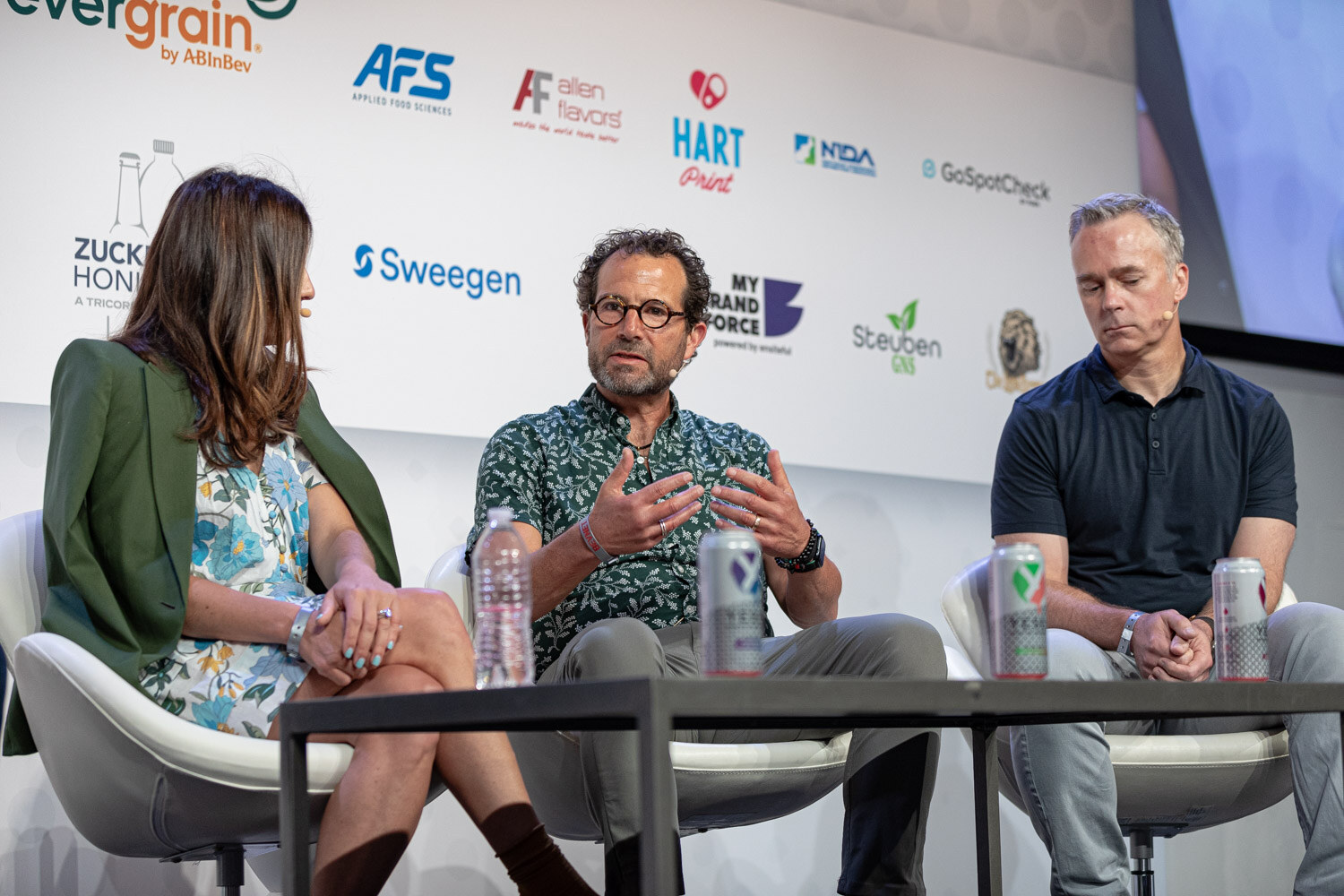 A woman in a green blazer and floral dress and two men in a patterned shirt and a navy polo sit on stage in white chairs with cans on the table.