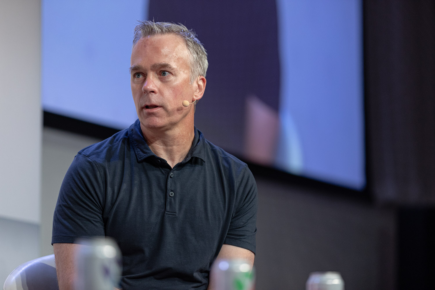 A man in a navy polo shirt looks to his left while seated on stage with an out-of-focus screen behind him.