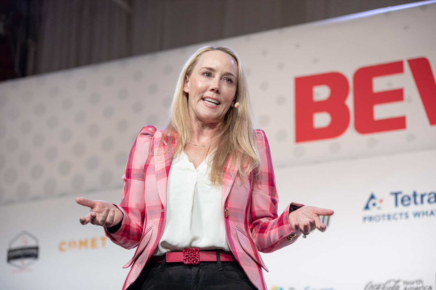A blonde woman in a pink plaid blazer and white top gestures with open hands in front of a large BevNET Live sign.