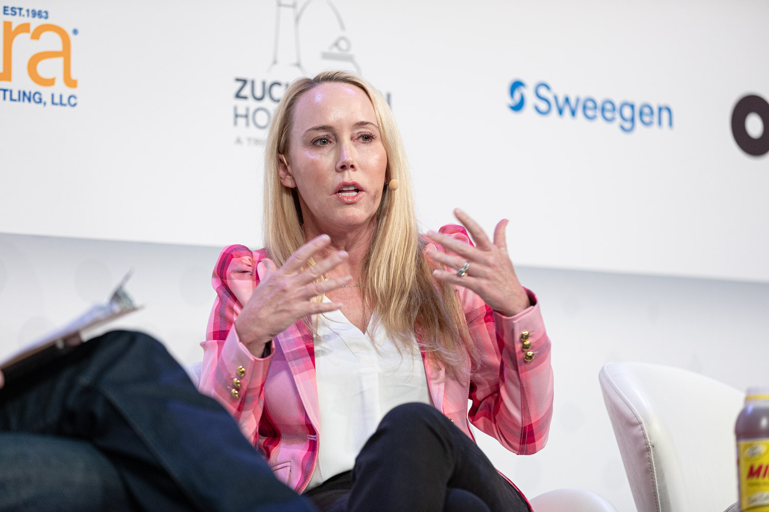 A blonde woman in a pink plaid blazer gestures with both hands while seated, with a Sweegen and Zuckerman Honickman sponsor wall behind her.