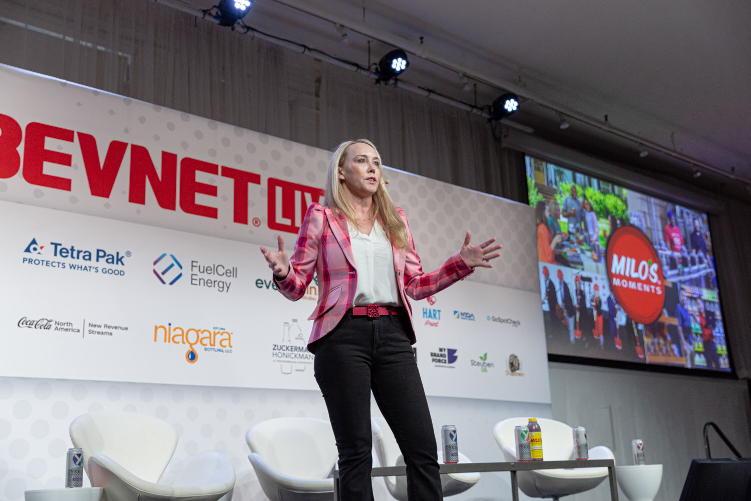 A woman in a pink blazer stands with arms open on a stage with white lounge chairs and a screen showing a Milo's Moments logo.