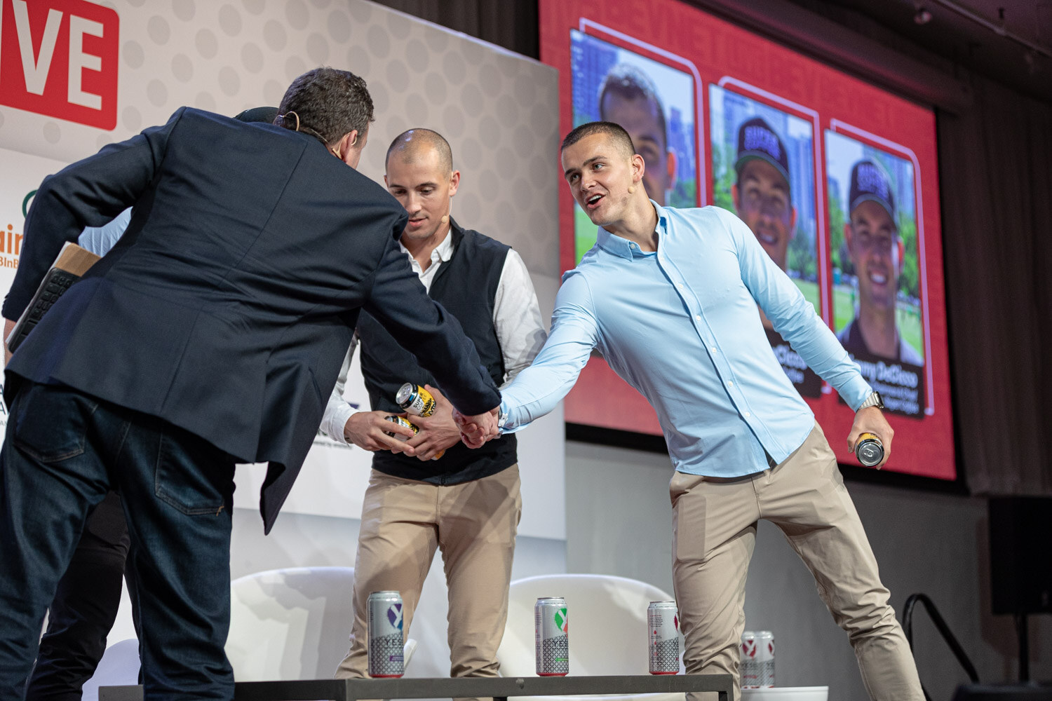 A man in a navy blazer shakes hands with two men in light blue and black vests as a screen with portraits glows behind them.