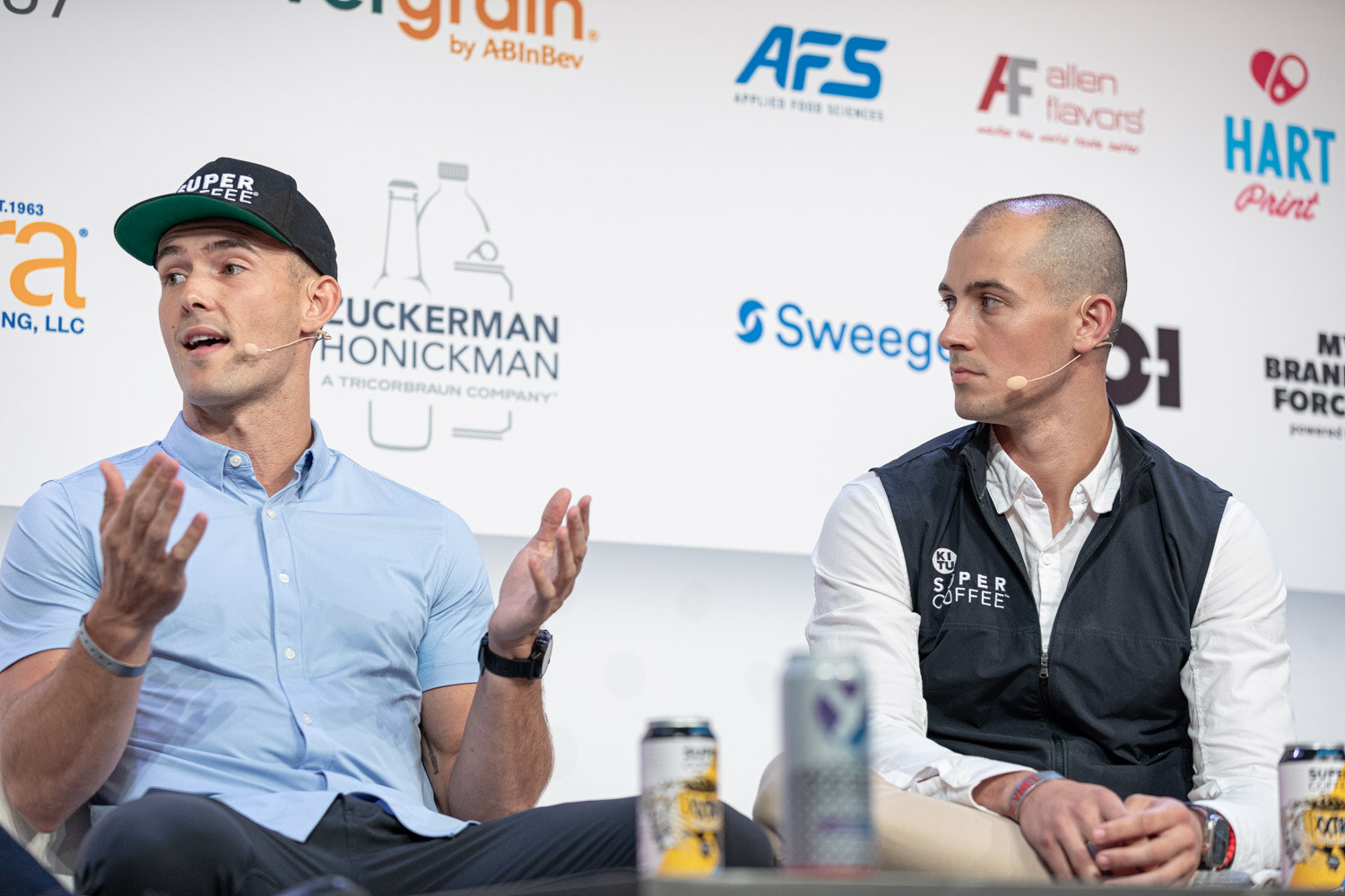 Two men seated on stage, one in a green and black Super Coffee cap and light blue shirt and one in a black vest with a Super Coffee logo, gesture while talking.