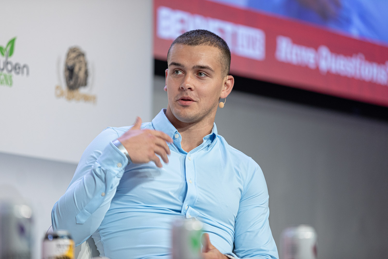 A young man in a light blue button-up shirt gestures with one hand while seated on stage in front of a white sponsor wall.