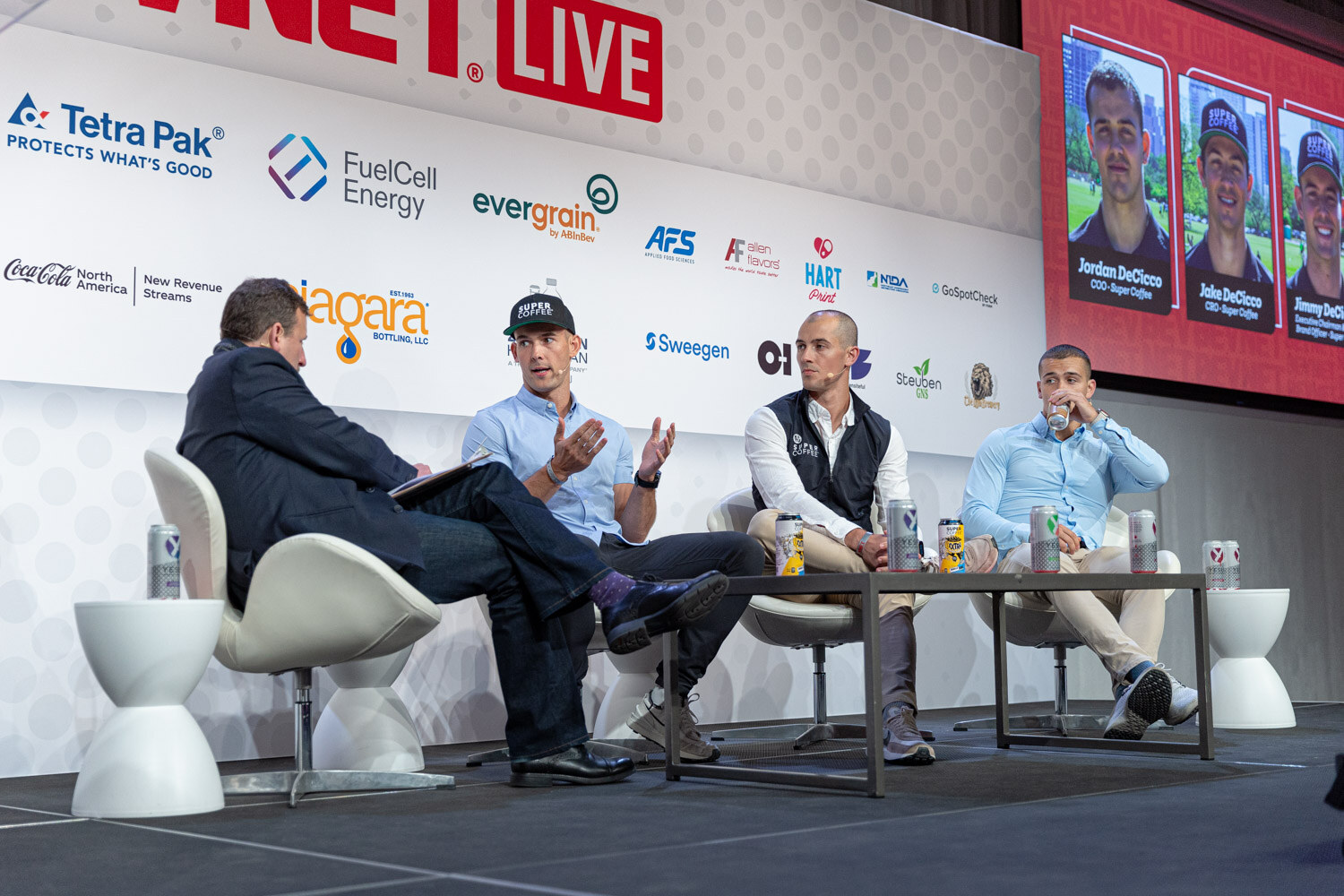 Wide shot of four people seated in white lounge chairs on a stage with a screen showing three portraits and sponsor logos.