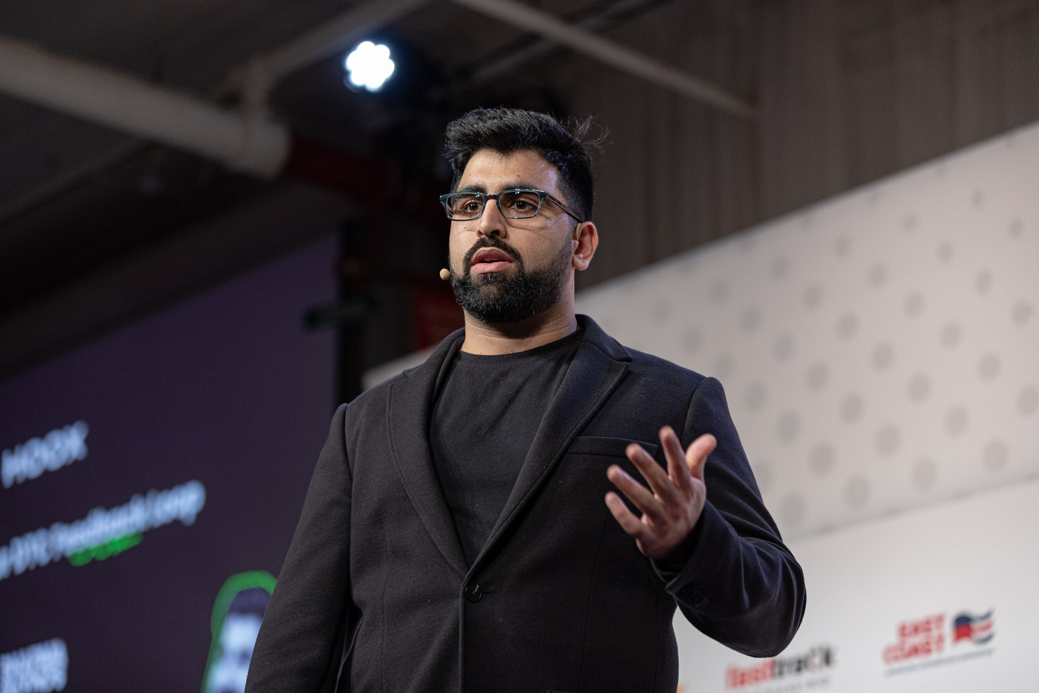 A bearded man with glasses in a dark jacket gestures while speaking under stage lights, with a dark slide behind him.