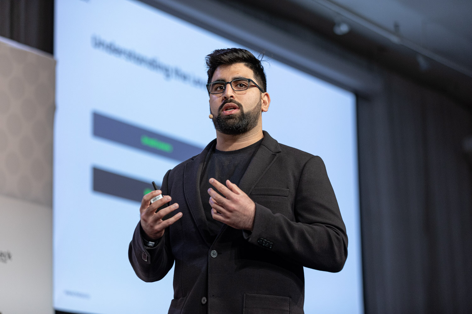 A man with glasses and a beard in a dark blazer holds a clicker in front of a large projected slide.
