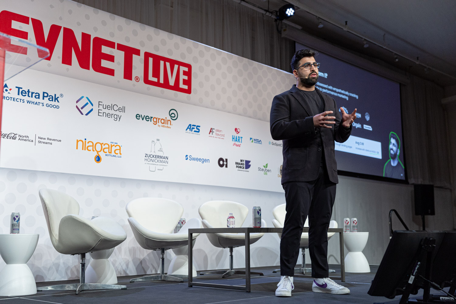 Wide shot of a man in dark clothes presenting on a stage with white lounge chairs and a BevNET Live backdrop with sponsor logos.