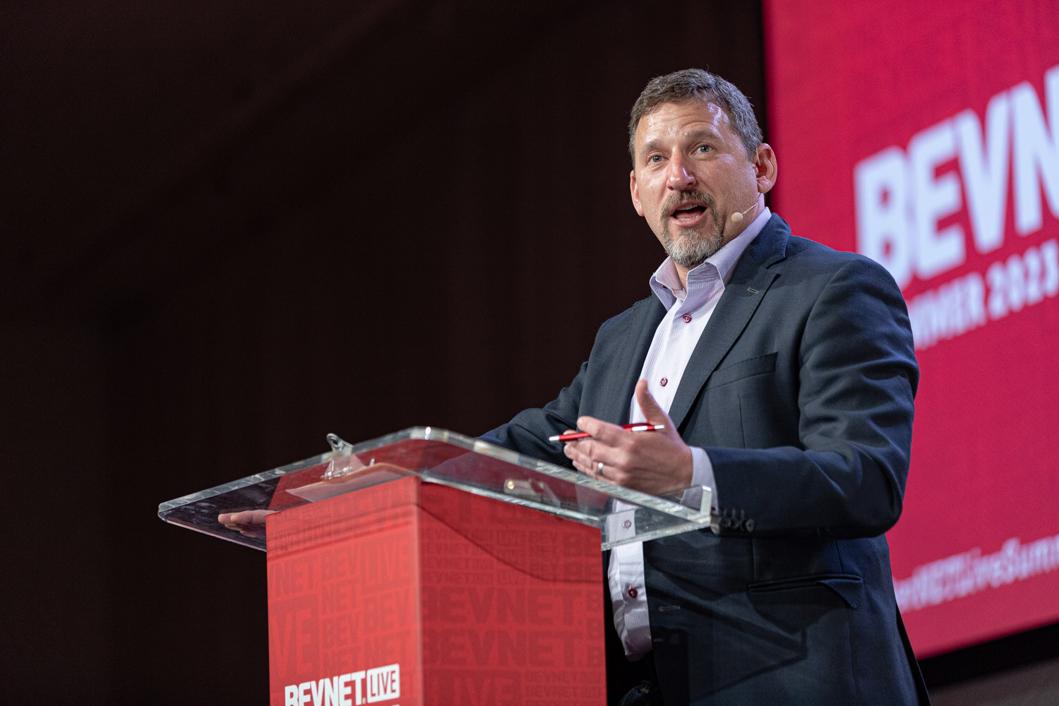 A bearded man in a navy blazer speaks at a clear podium in front of a red BevNET Summer 2023 screen.