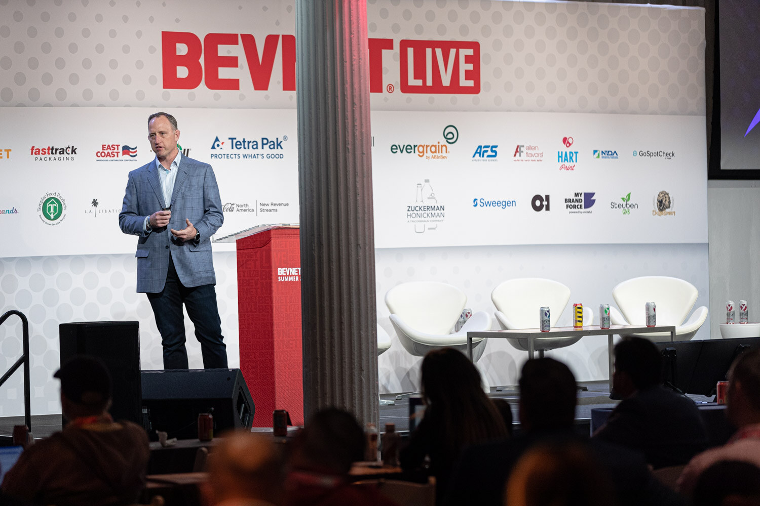 Wide shot of a man in a blue blazer standing on a stage next to a red podium with a lounge seating set and the BevNET Live backdrop behind him.