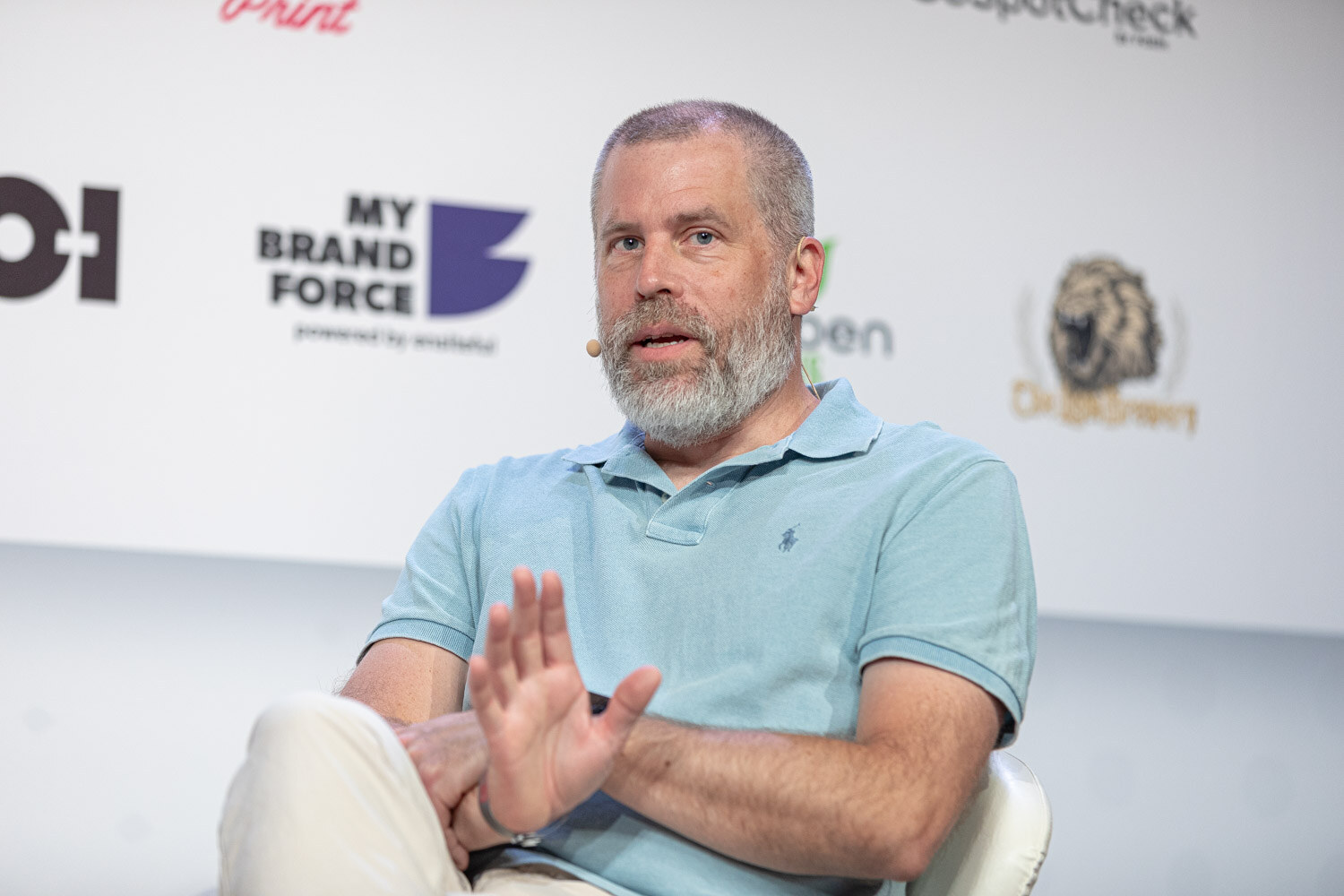 A man with a gray beard in a light blue polo shirt holds a hand up while speaking on stage in front of a sponsor wall.