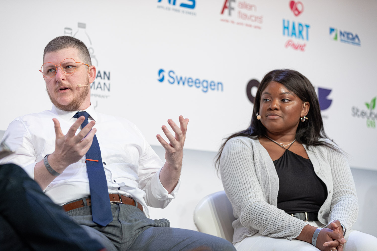 A man in a white shirt and navy tie gestures with both hands next to a woman in a gray cardigan and black top, seated on stage.