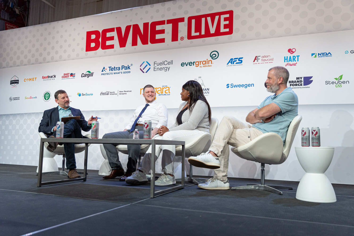 Wide shot of four people seated in white chairs around a low table on a stage with the BevNET Live logo on the wall behind them.