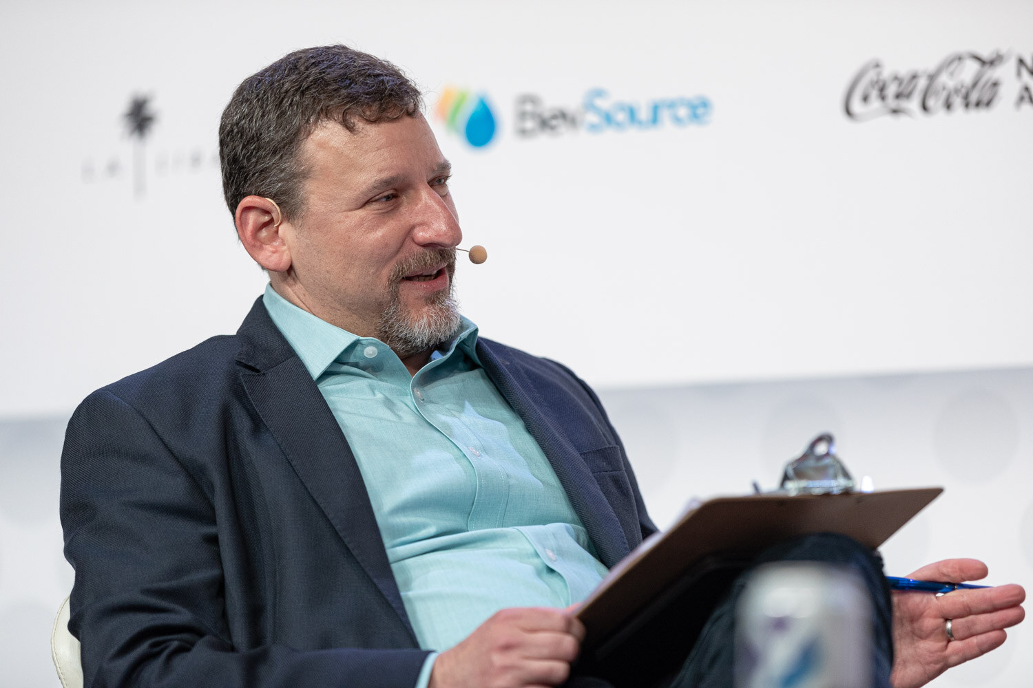 A bearded man in a navy blazer and teal shirt holds a clipboard and a pen while seated on stage with BevSource and Coca-Cola logos behind him.