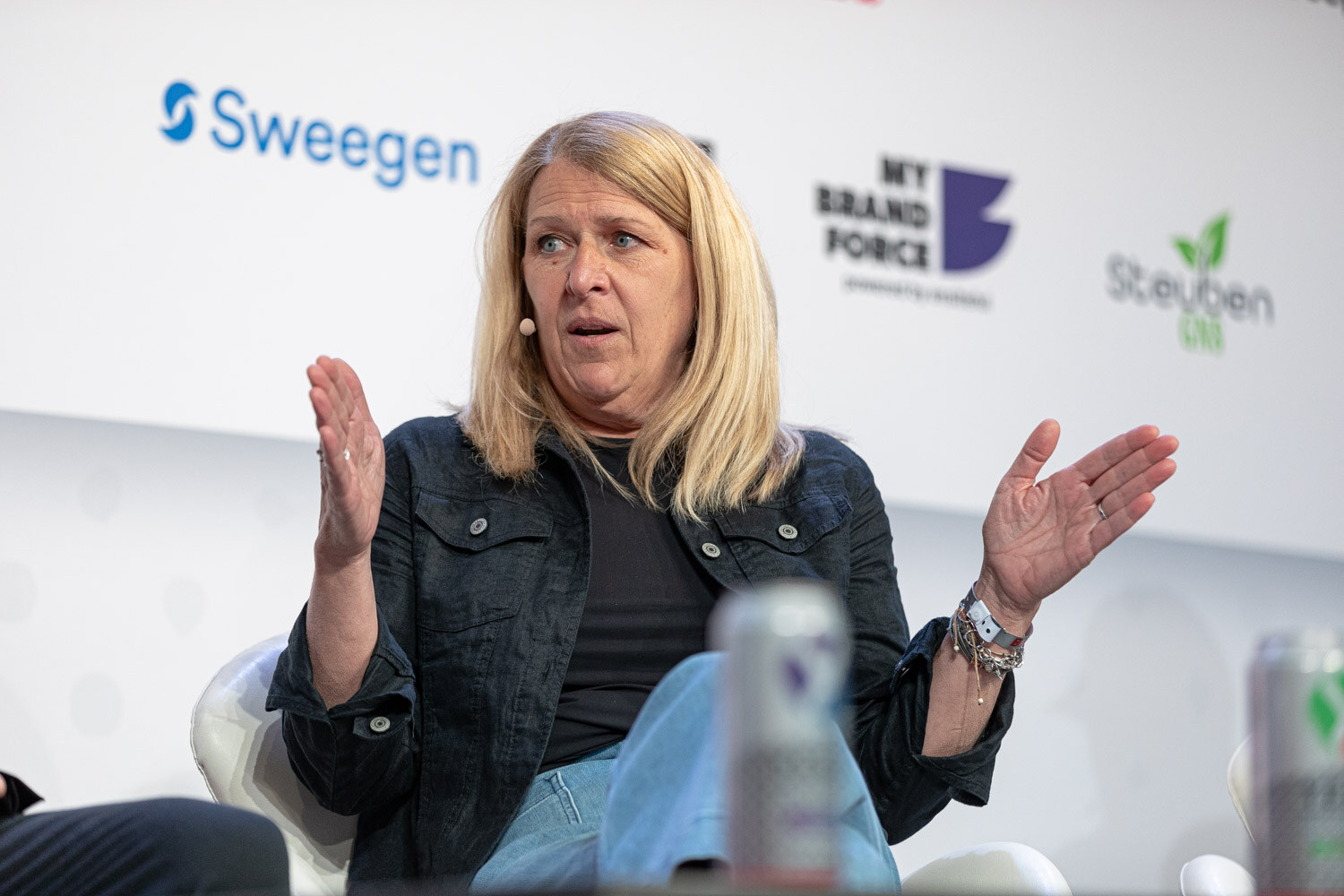 A blonde woman in a black denim jacket and light jeans gestures with open palms while seated on stage.