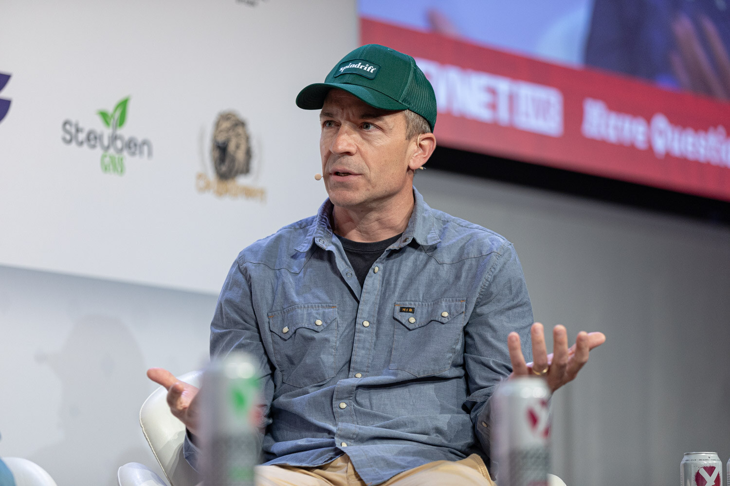 A man in a green cap and a blue snap-button shirt gestures with open hands while seated on stage with cans in the foreground.