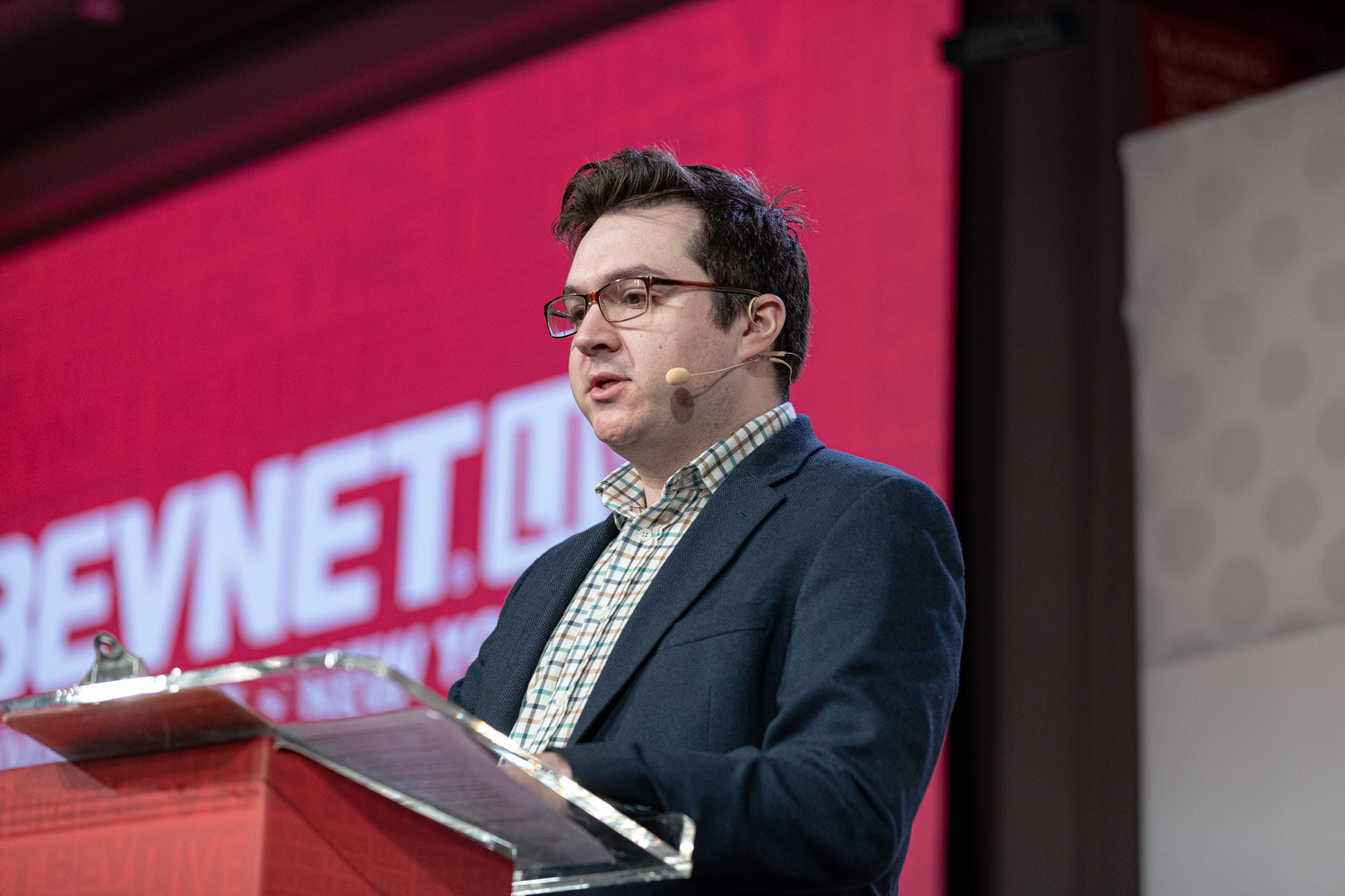A man with glasses in a navy blazer and checked shirt speaks into a headset microphone at a clear podium in front of a red screen.