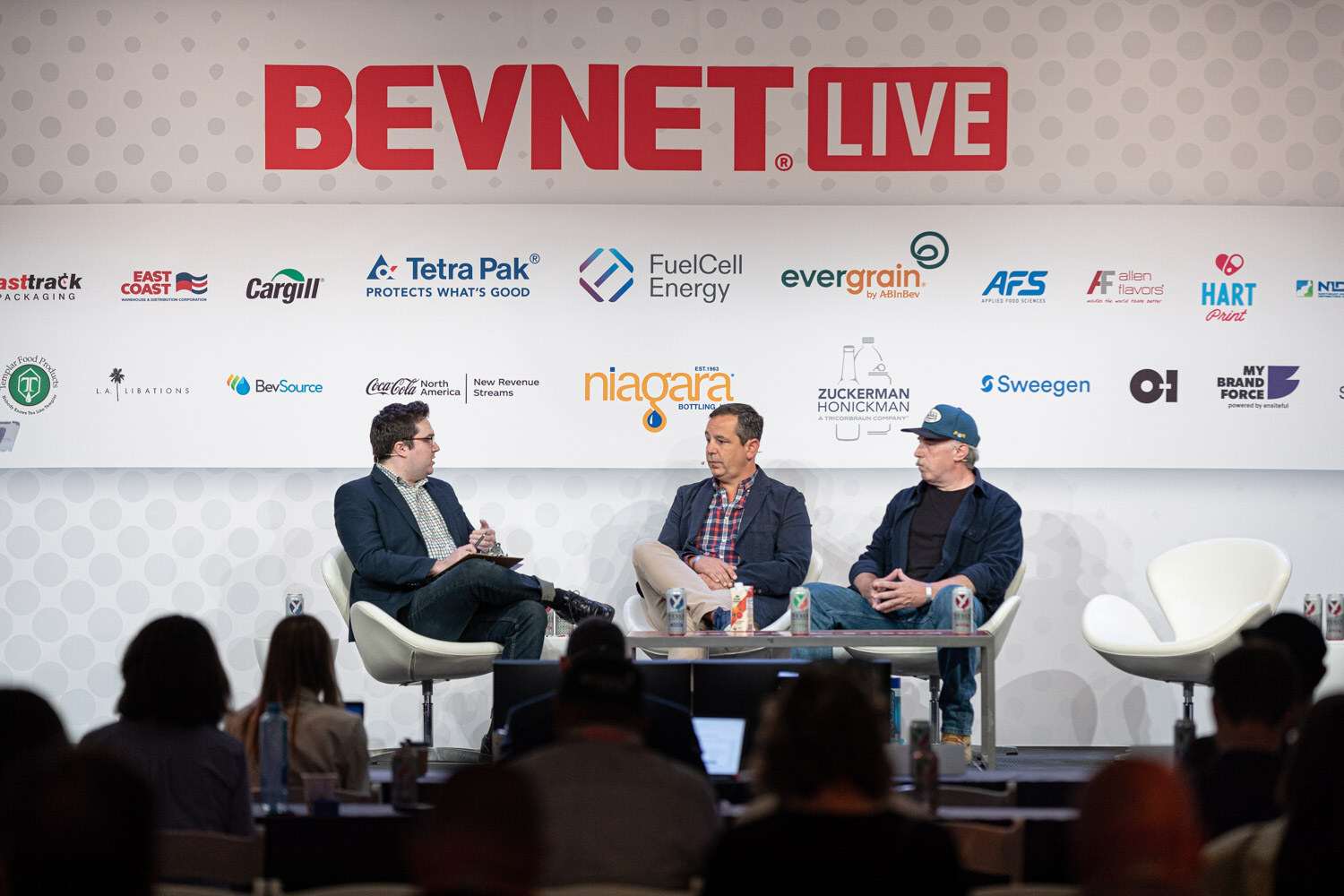 Wide shot of three men seated in lounge chairs on a stage under the BevNET Live logo with a silhouetted audience in front.