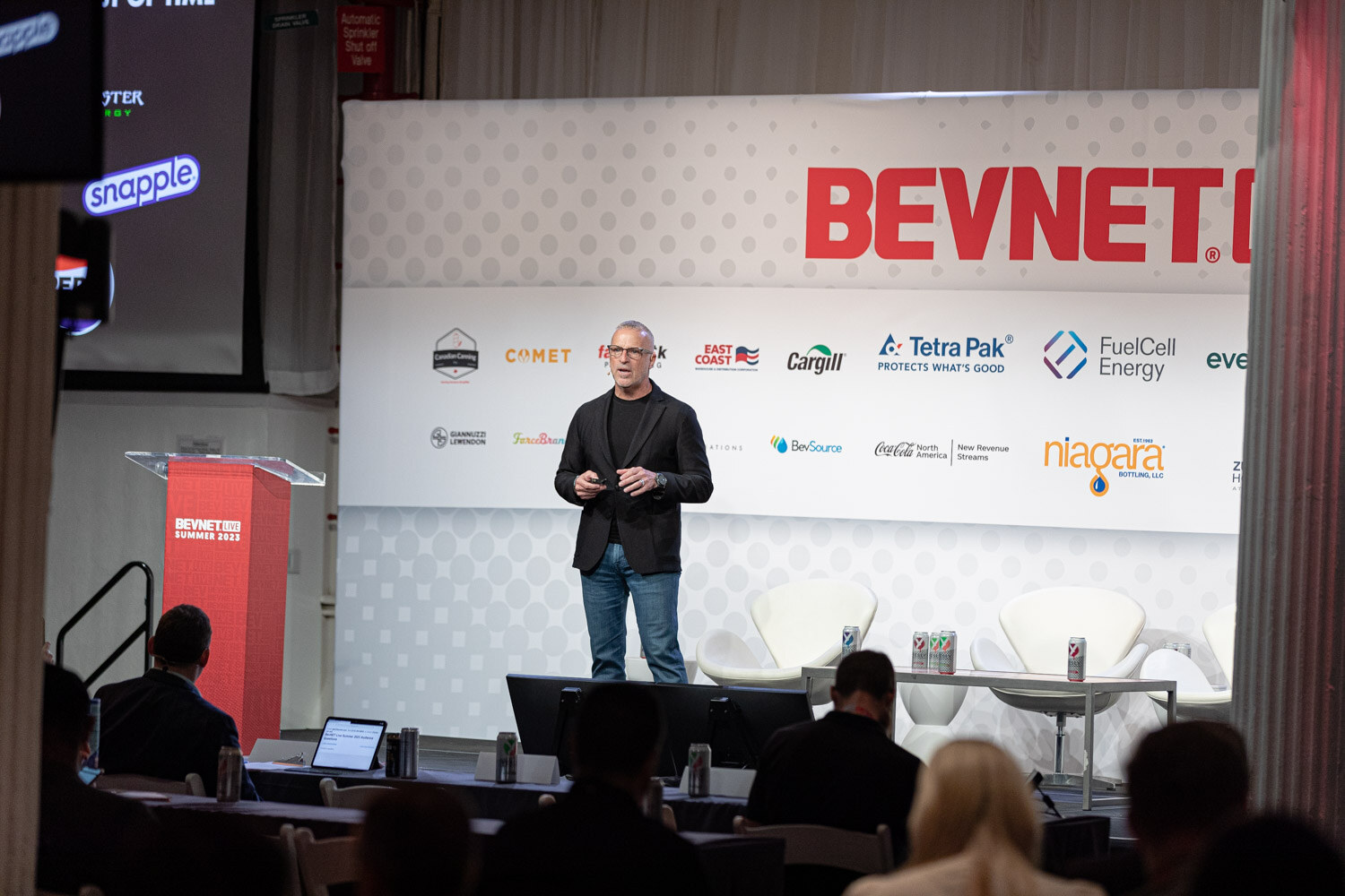 Wide shot of a man in a dark blazer and jeans standing on a stage beside a red podium with the BevNET Live backdrop and an audience in the foreground.