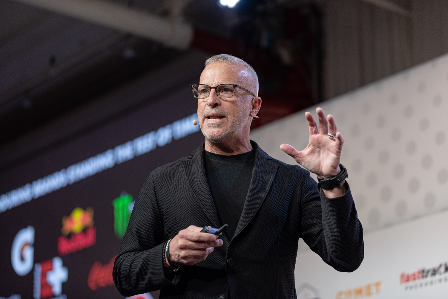 A gray-haired man with glasses in a black blazer holds a clicker and raises one hand in front of a dark slide with beverage brand logos including Gatorade and Red Bull.