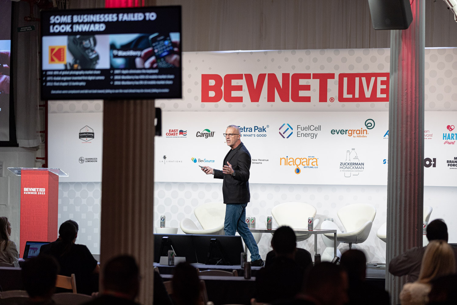 A man in dark clothes walks across a stage in front of the BevNET Live backdrop with a monitor at upper left showing a slide that reads Some businesses failed to look inward.