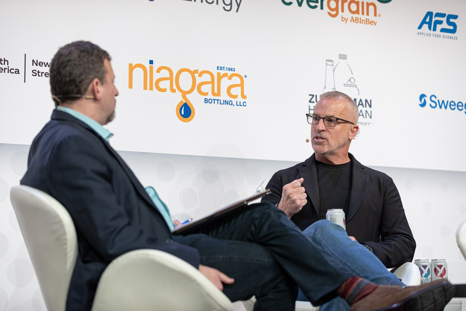 A man in a navy blazer with a notepad sits facing a gray-haired man with glasses in a black blazer, both seated on stage in front of a Niagara sign.