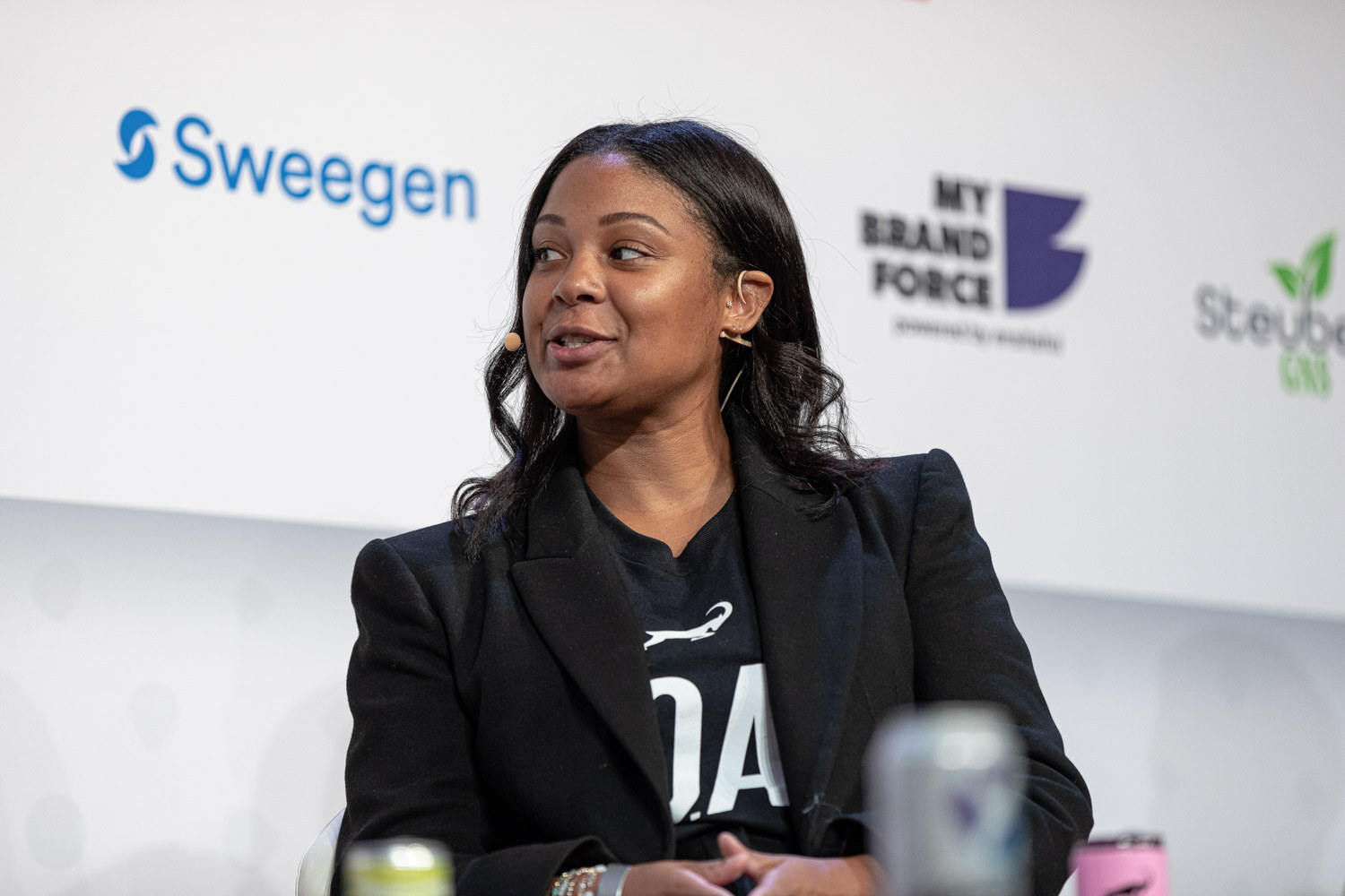 A woman in a black blazer over a dark top with a white logo speaks with a Sweegen and My Brand Force sponsor wall behind her.