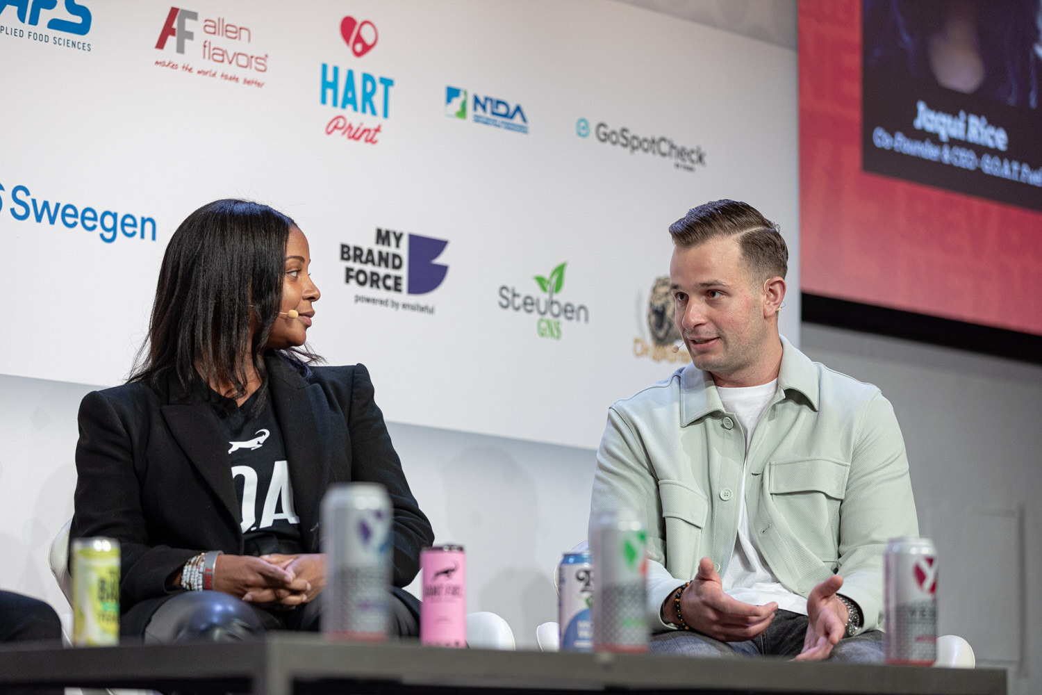 A woman in a black blazer and a man in a light green shirt jacket sit on stage in front of a screen with a name caption, with colorful cans on the table in front of them.
