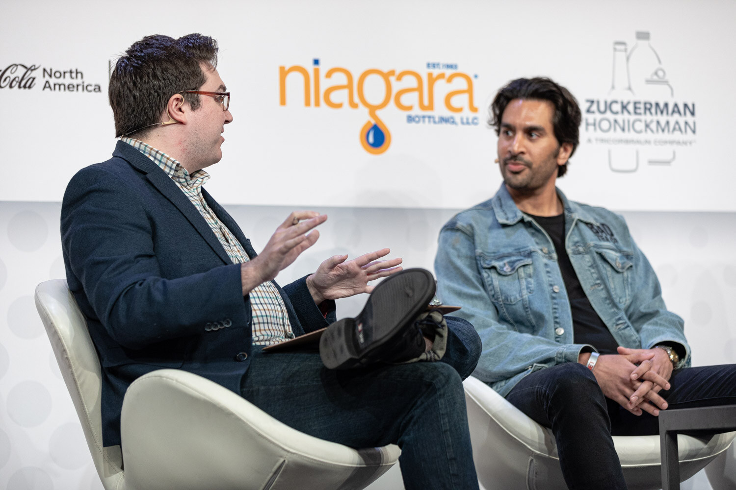A man in a dark blazer with a clipboard gestures toward a man in a blue denim jacket seated beside him in front of a Niagara and Zuckerman Honickman sign.