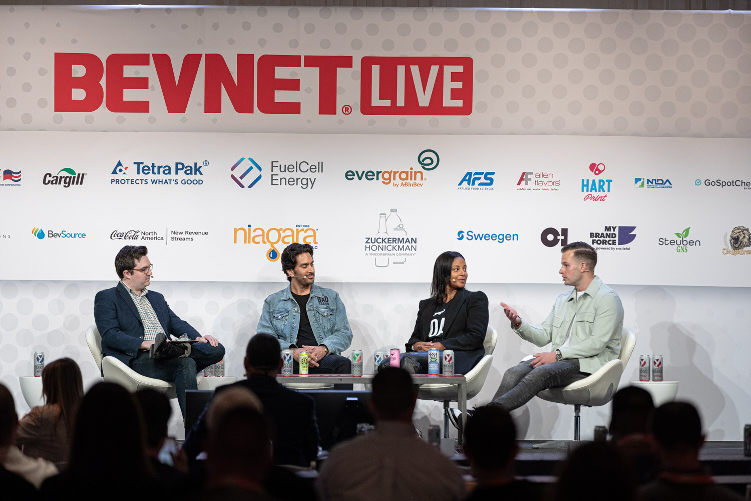 Wide shot of four people seated in white chairs on a stage with the BevNET Live logo above them and cans on the table.