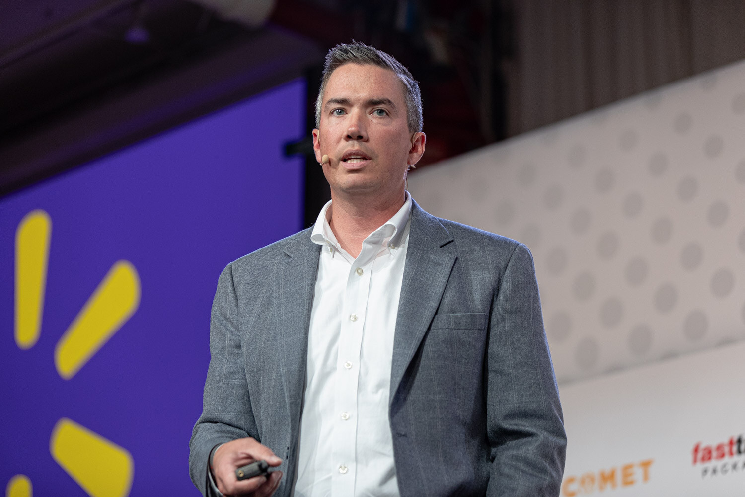 A man in a gray blazer and white shirt holds a presentation clicker in front of a purple slide with a yellow Walmart spark logo.