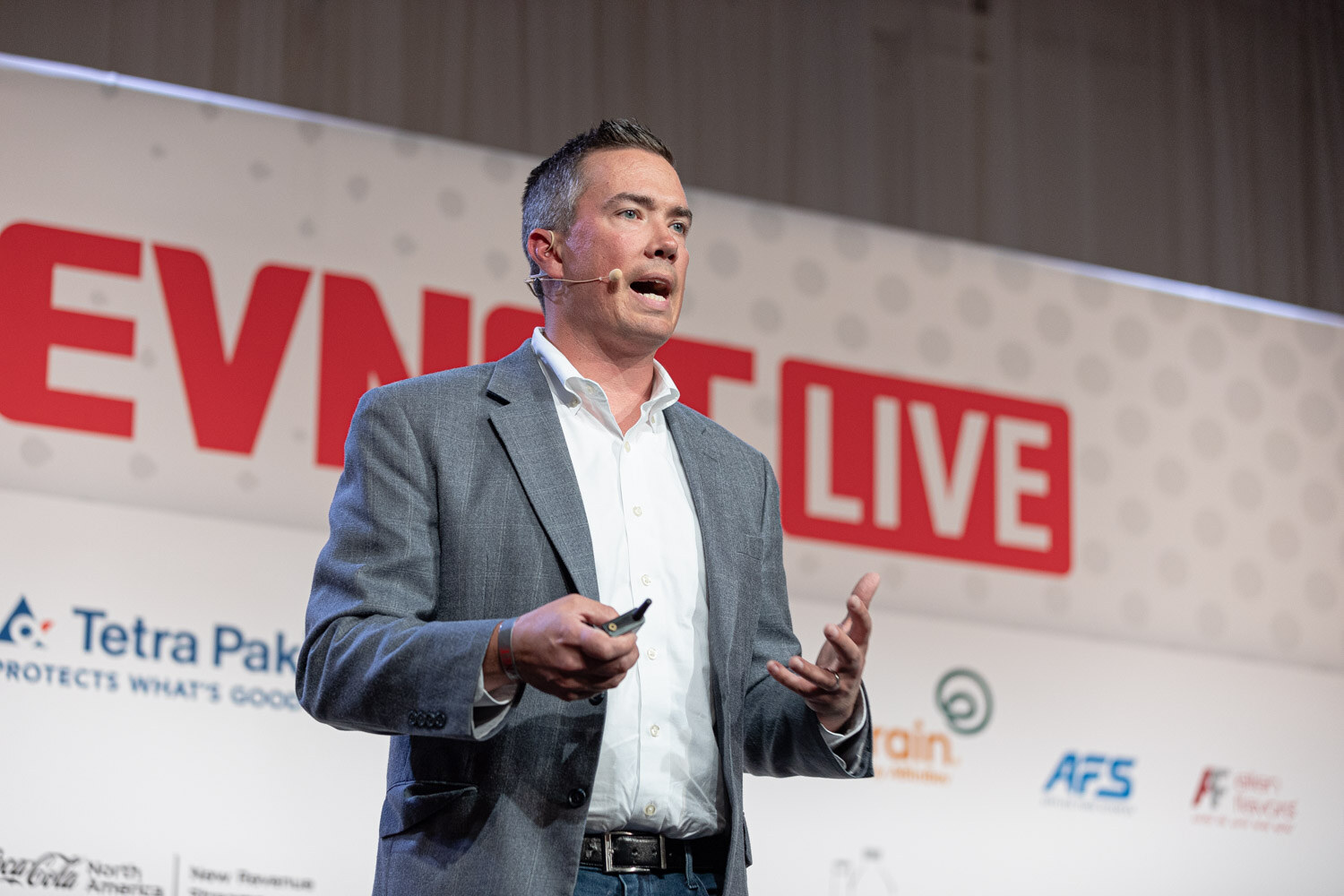 A man in a gray blazer speaks with open hands in front of a large BevNET Live sign on the stage backdrop.