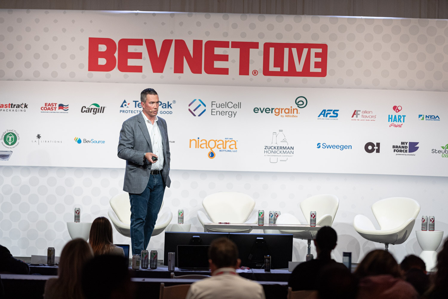 Wide shot of a man in a gray blazer and jeans standing on a stage in front of the BevNET Live backdrop with white lounge chairs behind him and audience heads in the foreground.