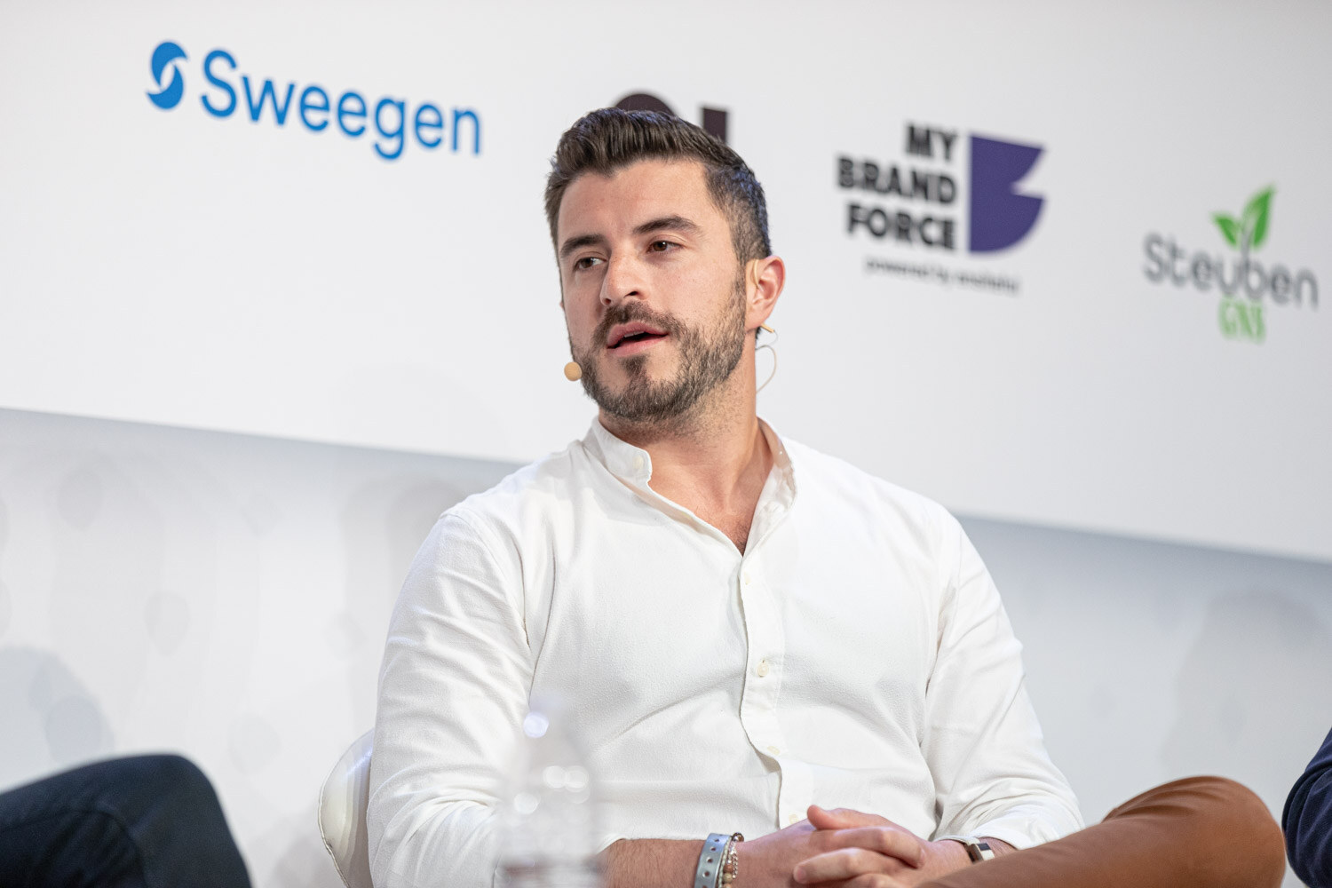 A man with a trimmed beard in a white button-up shirt looks to his left while seated on stage in front of a Sweegen sponsor sign.