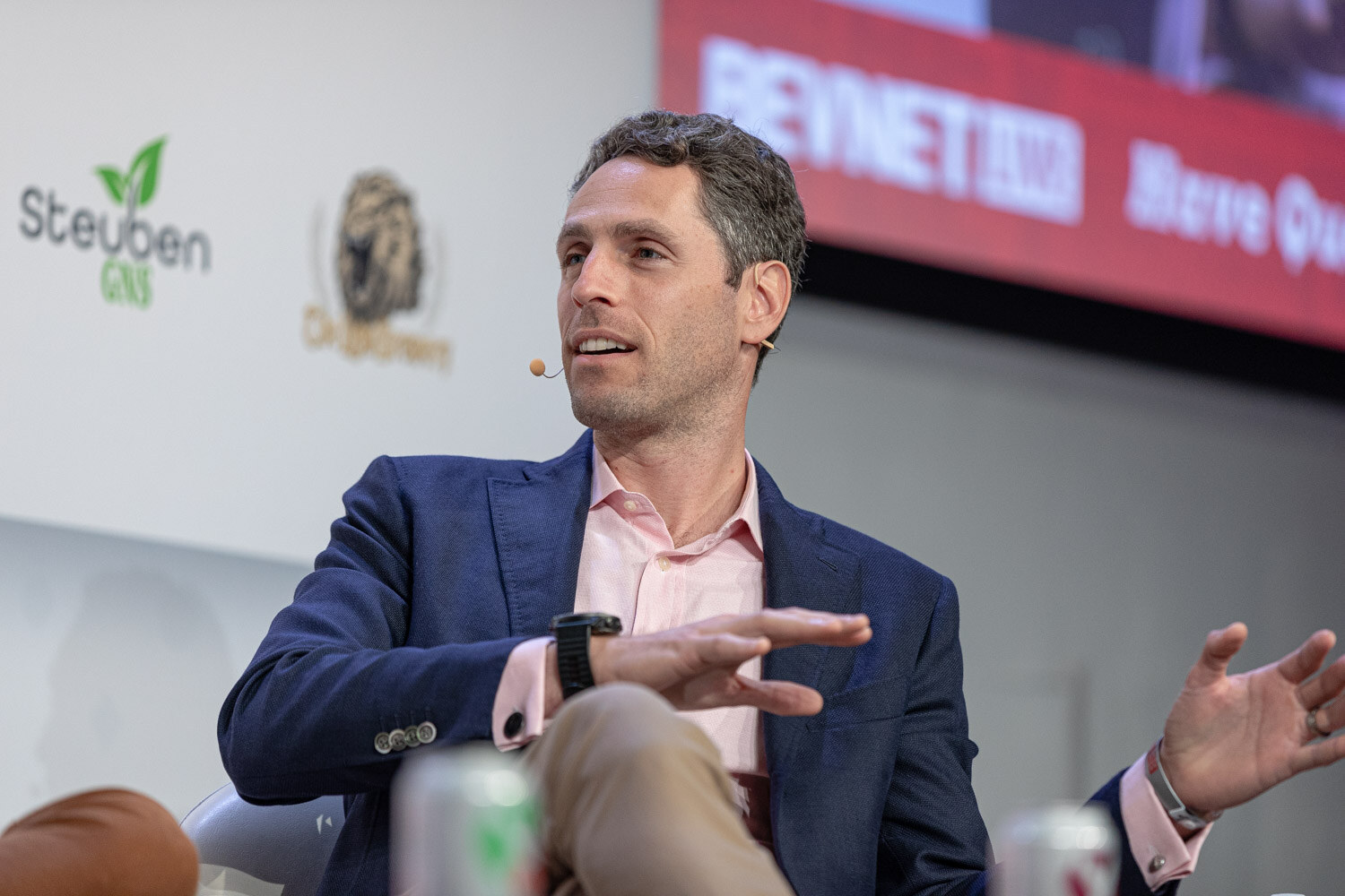 A man in a navy blazer and pink collared shirt gestures with one hand while seated on stage with a red screen behind him.