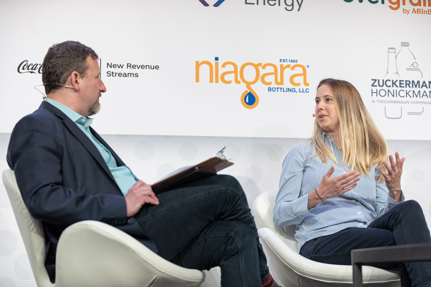A man in a dark blazer holding a clipboard faces a blonde woman in a light blue striped shirt, seated in white chairs in front of a Niagara sign.