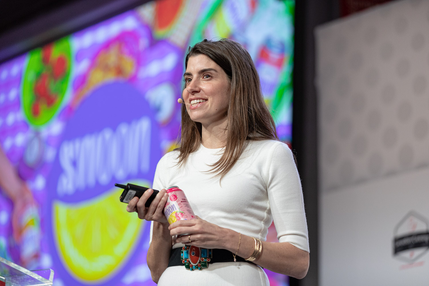 A woman in a white top and black belt smiles while holding a presentation clicker and a pink can in front of a large purple and green slide reading Swoon.