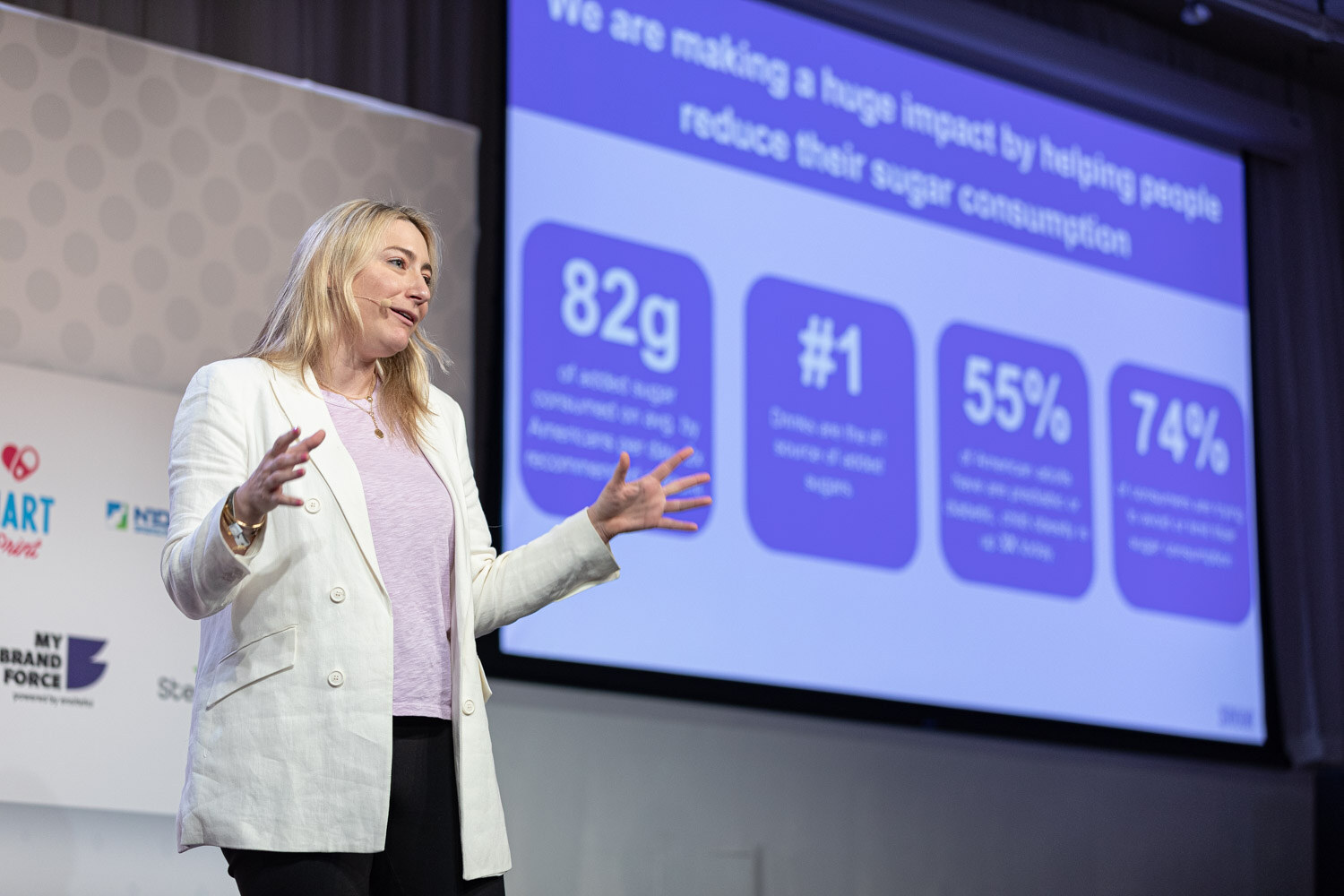 A blonde woman in a white blazer gestures on stage next to a projected slide with statistics including 82g, number 1, 55 percent and 74 percent.