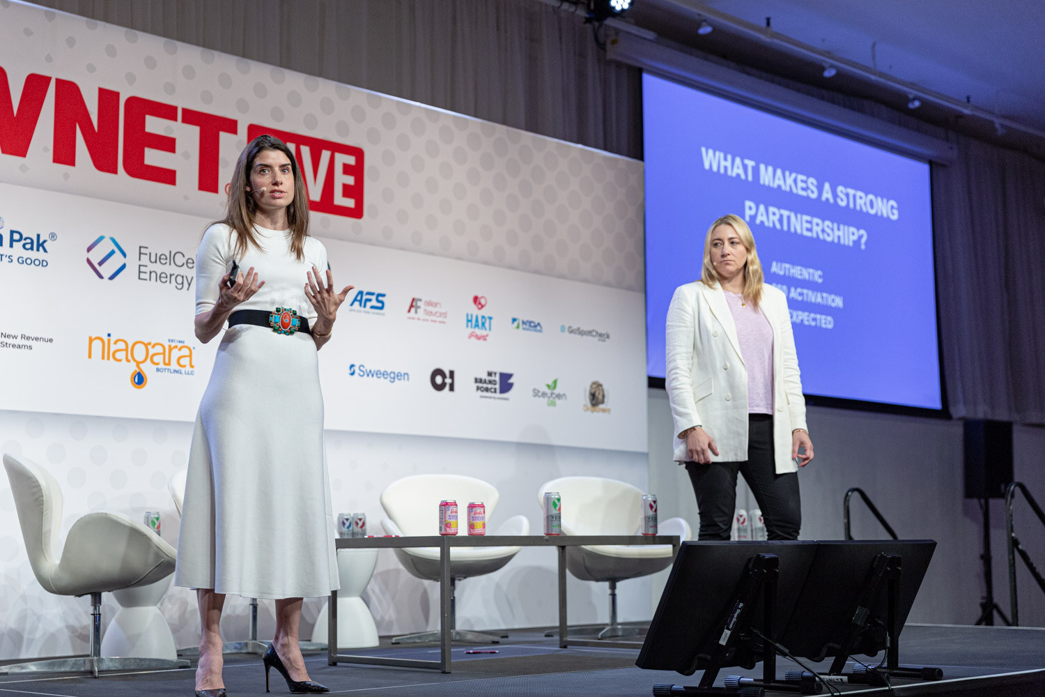 Wide shot of two women in white outfits on a stage with white lounge chairs and a blue slide reading What Makes a Strong Partnership.