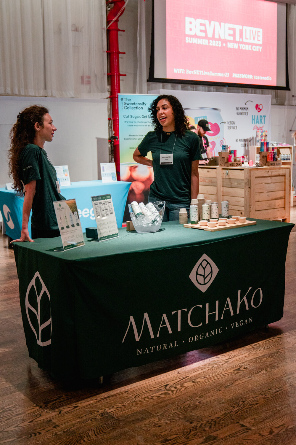 Two women stand behind a dark green table with the Matchako logo and small product displays on a wooden floor.
