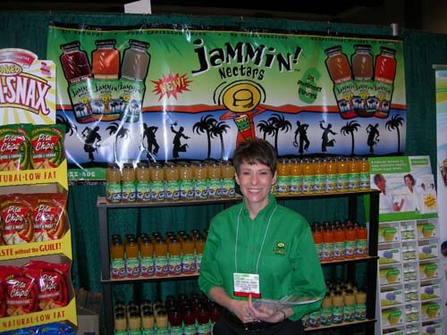 A smiling woman with short dark hair in a green shirt and badge stands before shelves of bottled drinks under a Jammin' Nectars banner with palm tree silhouettes.