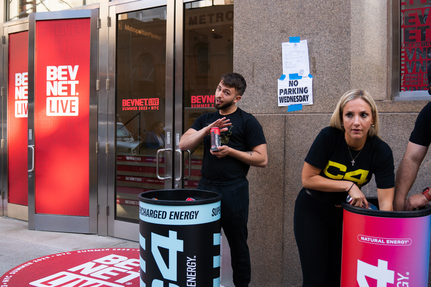 A man in a black T-shirt and a woman in a black C4 shirt stand beside a C4 Energy cooler at the red BevNET Live doors on the sidewalk.