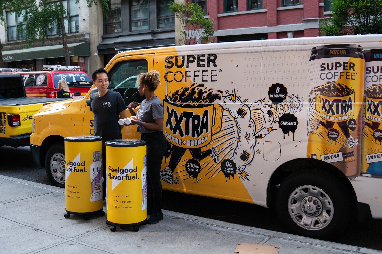 Staff in dark shirts stand beside two yellow Feelgood flavorfuel coolers in front of a yellow Super Coffee XXTRA van on the street.