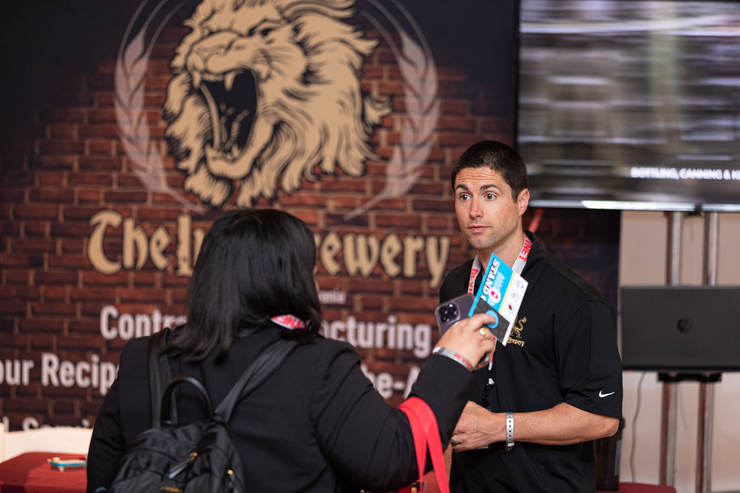 A man in a black polo and a woman with a backpack talk in front of a brick-style booth wall showing a gold lion logo and the words Contract Manufacturing.