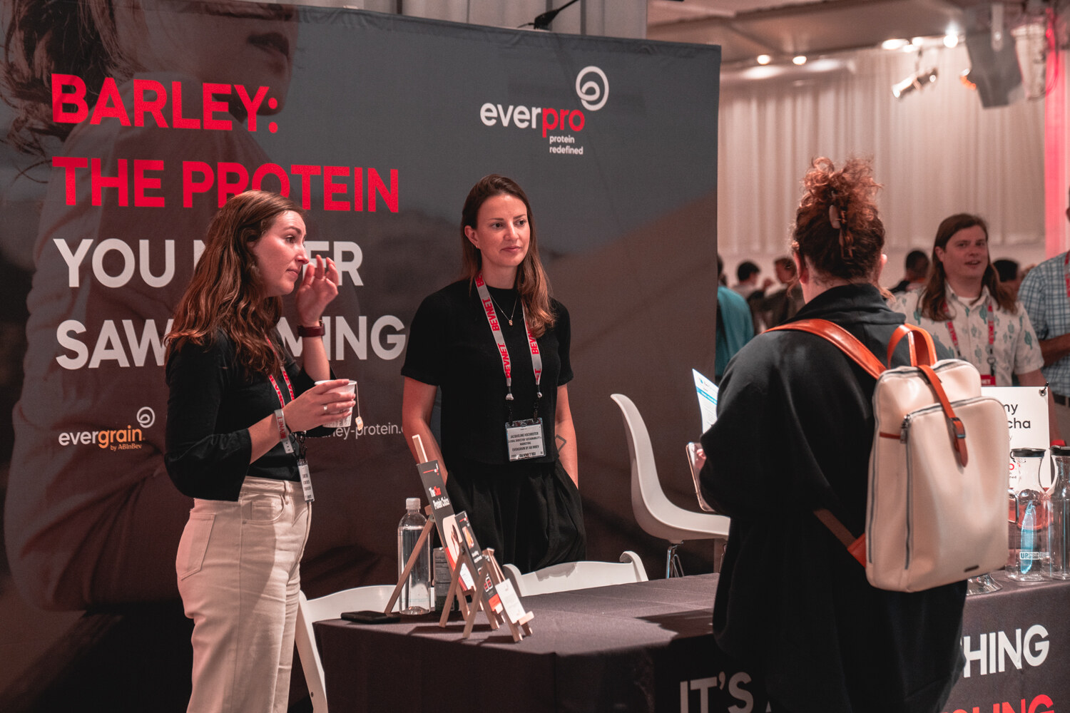 Two women in front of a dark booth with a red headline about barley and protein talk while an attendee with a backpack looks on.