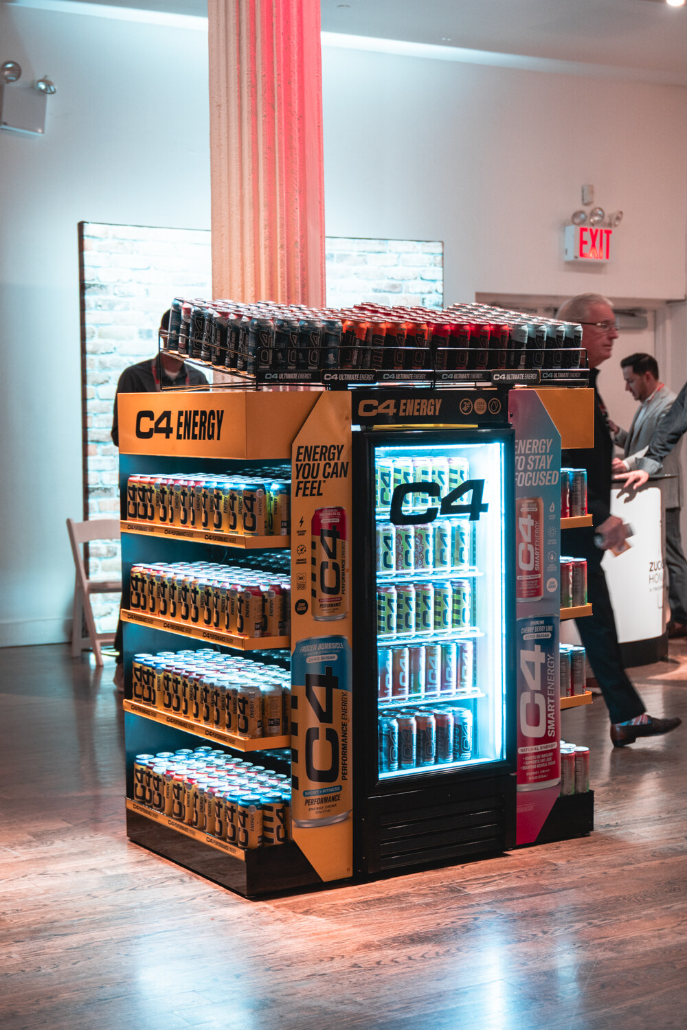 A lit freestanding C4 Energy display with shelves of cans and a glass-door cooler stands beside a pink-lit column.