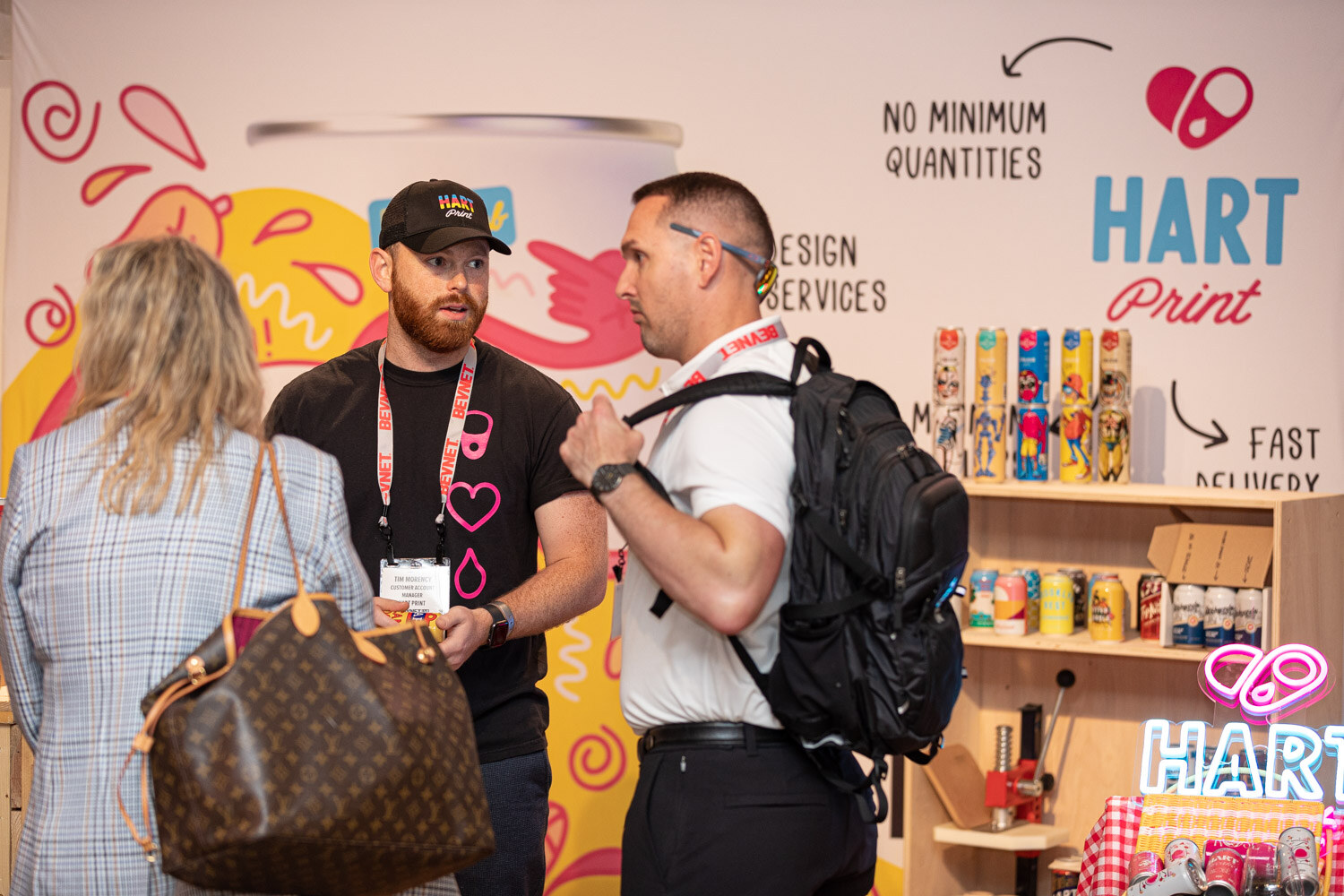 A bearded man in a Hart Print cap and another man with a backpack talk to a woman at a booth with a colorful mural and the Hart Print logo.