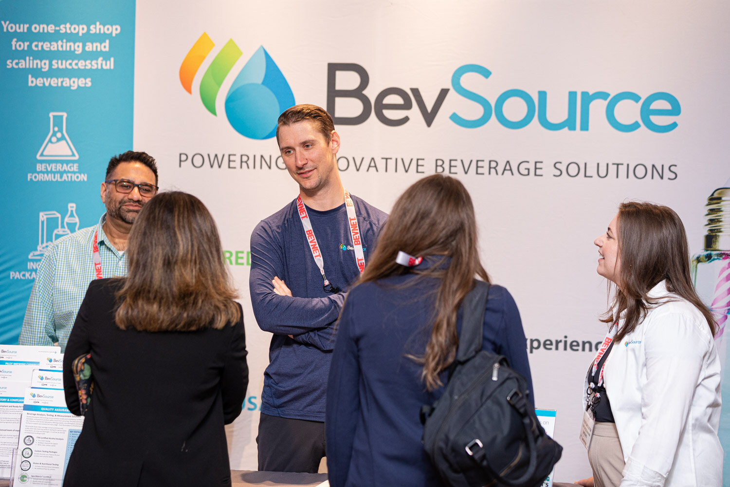 Two men in front of a booth banner reading BevSource, Powering Innovative Beverage Solutions, speak with two women with their backs to the camera.