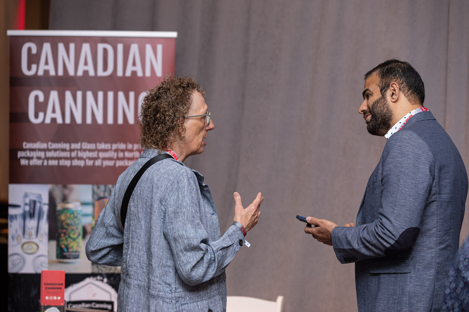 A woman in a gray jacket and a man in a blazer talk in front of a sign reading Canadian Canning.